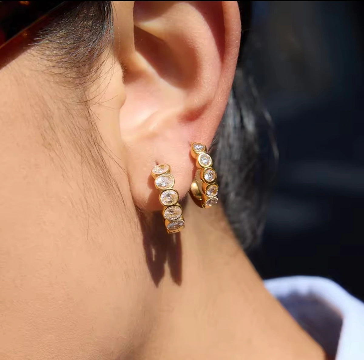 A close-up of a person's ear wearing two gold huggie hoop earrings, each set with multiple clear round gemstones.