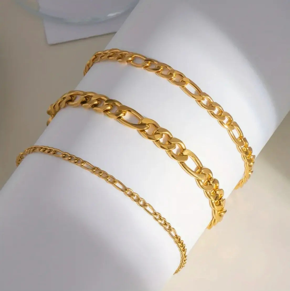 Three gold-toned chain bracelets displayed on a white cylindrical mannequin arm against a light beige background.