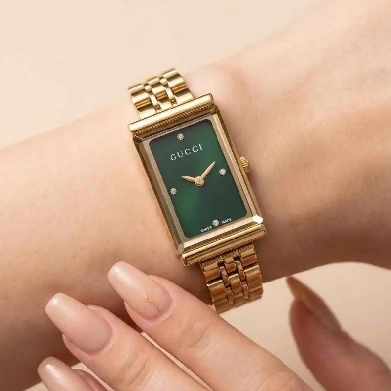 A close-up of a Gucci women's watch with a gold-tone rectangular case and link bracelet, featuring a green dial with diamond hour markers, worn on a wrist.