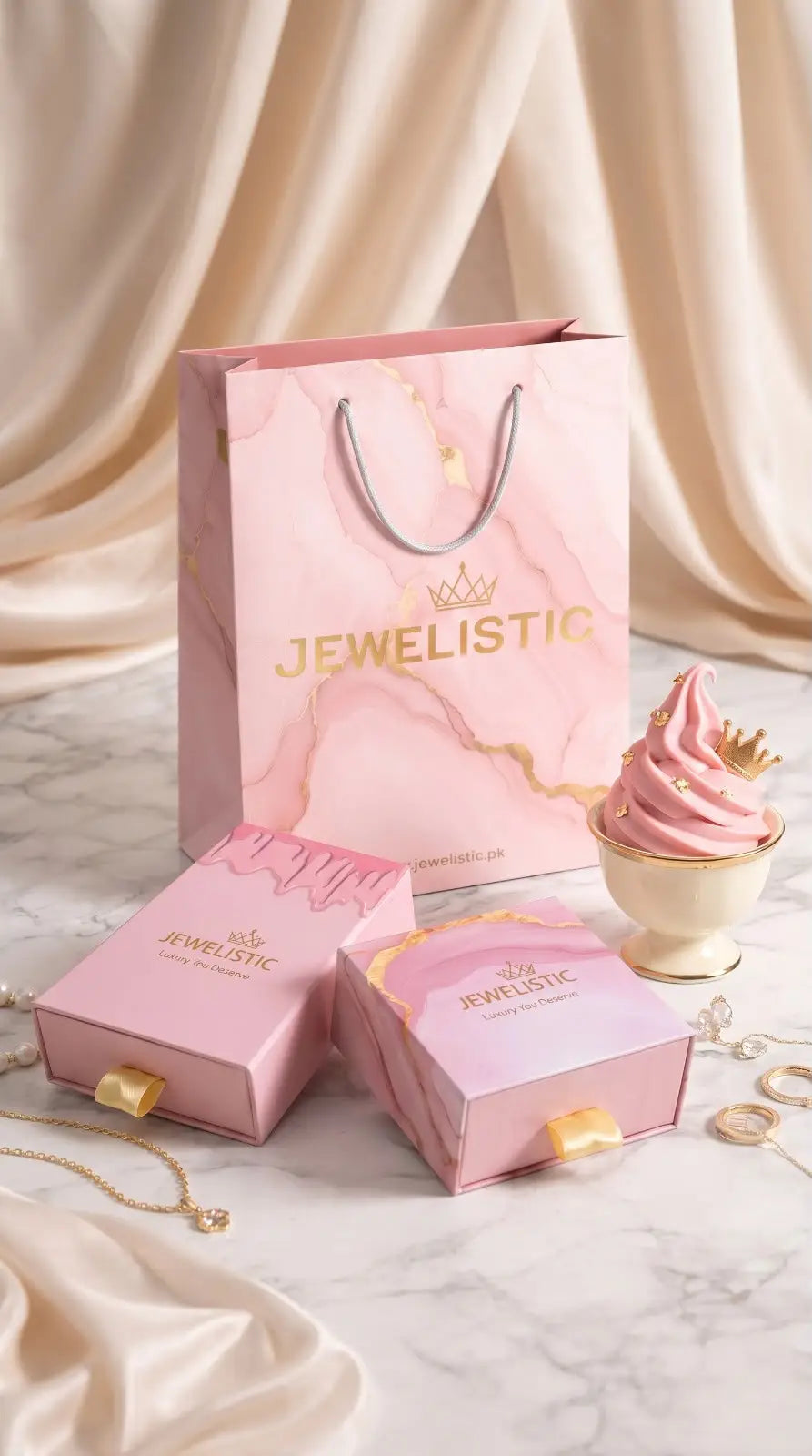 Pink 'Jewelistic' jewelry box, necklace, and cupcake on a marble surface with draped fabric background
