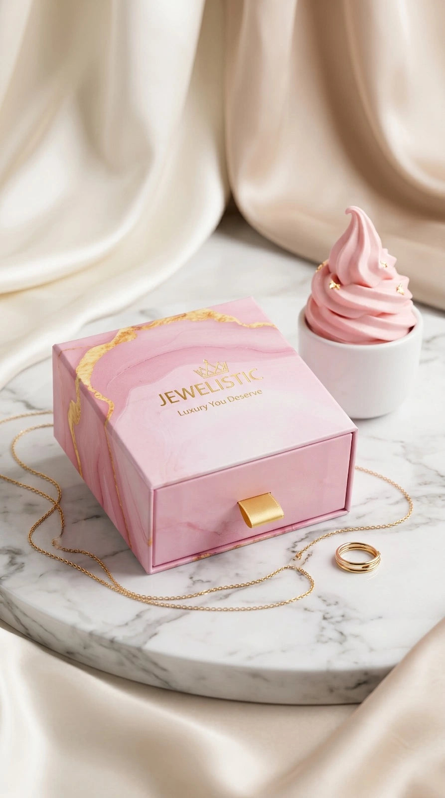 Pink jewelry box with gold accents on a marble surface, next to a pink dessert and gold necklace.