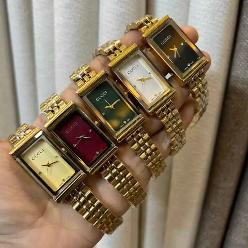 Four Gucci women's analog watches with rectangular gold-tone cases and metal link bracelets, held in a hand against a beige curtain background. The watch faces are shown in gold, red, green, and black colorways.