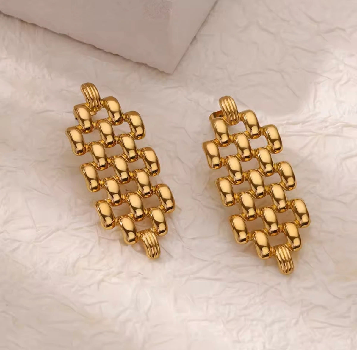 A pair of gold-toned rectangular link earrings with a textured, beaded chain-like design, displayed on a white textured surface.