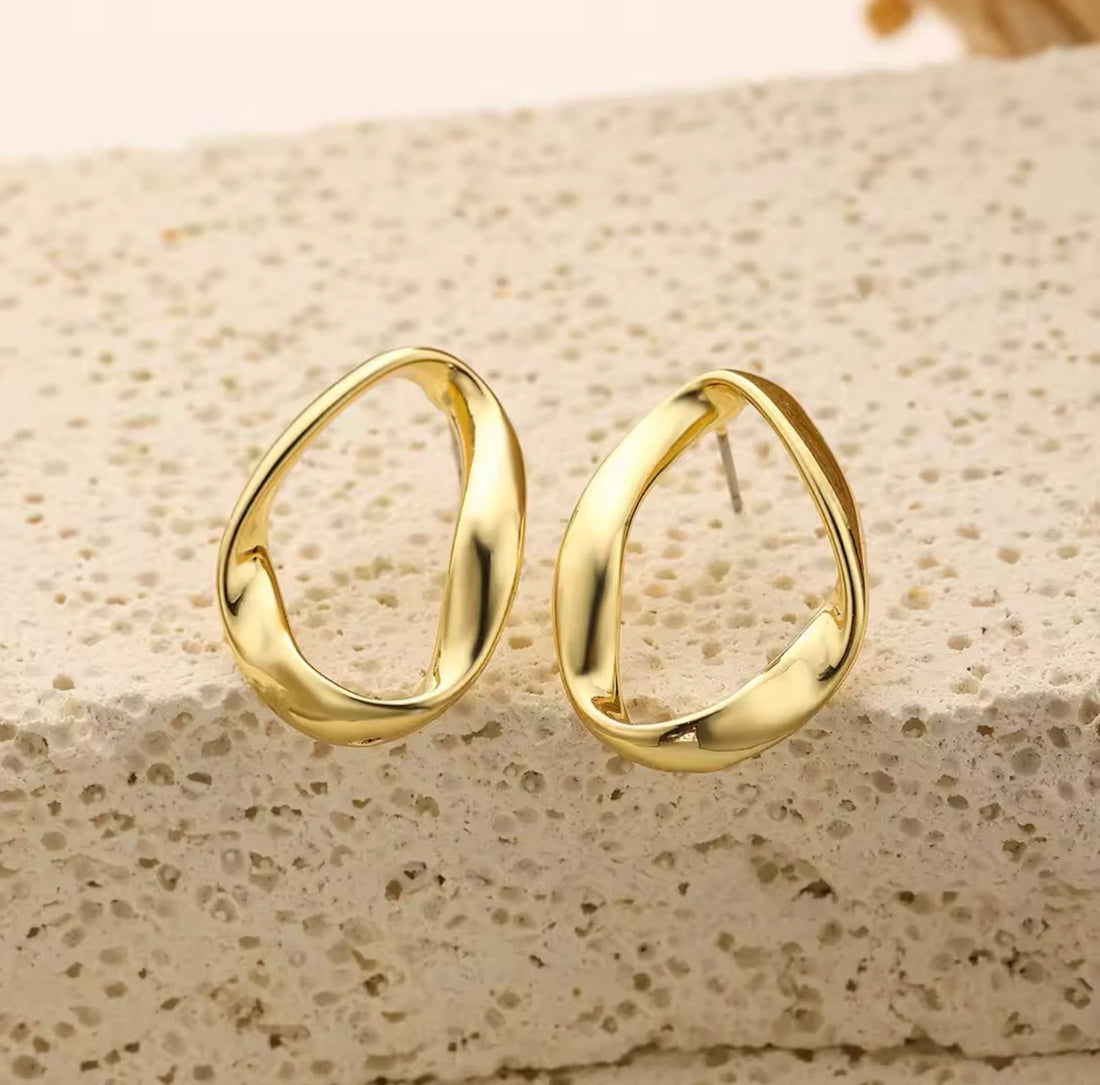A pair of gold-toned abstract open hoop earrings displayed on a textured beige stone surface.
