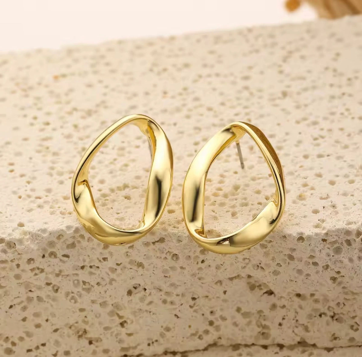 A pair of gold-toned abstract open hoop earrings displayed on a textured beige stone surface.