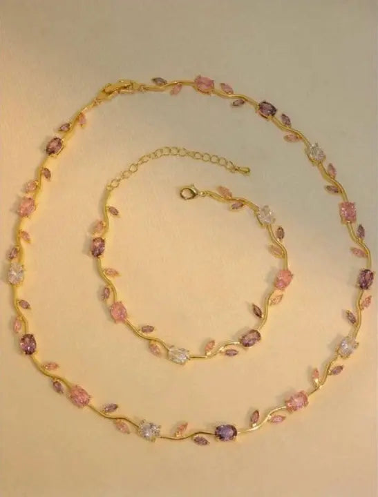 Gold floral vine necklace and matching bracelet set with pink and purple gemstone accents on a beige background.