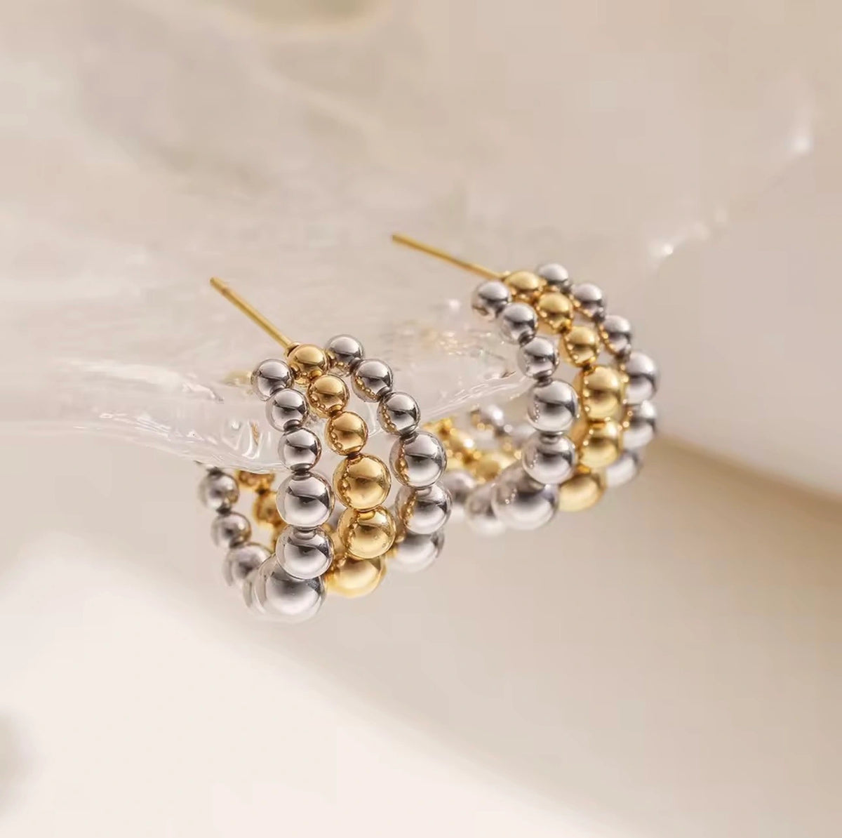 Two gold and silver beaded hoop earrings resting on a clear glass surface against a soft, warm off-white background.