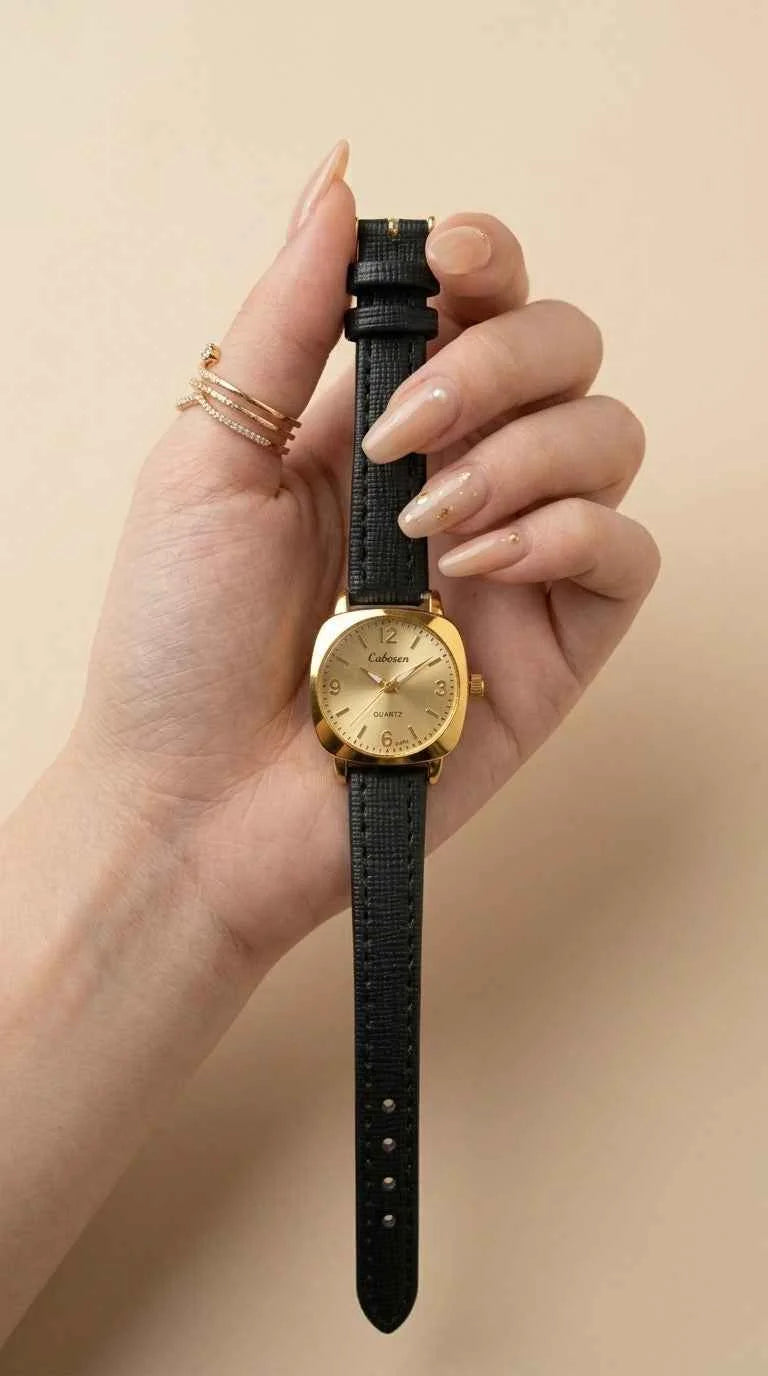A close-up of a woman's wrist and hand wearing a vintage-style watch with a gold-toned case, gold dial, and dark brown leather strap, set against a soft beige background.
