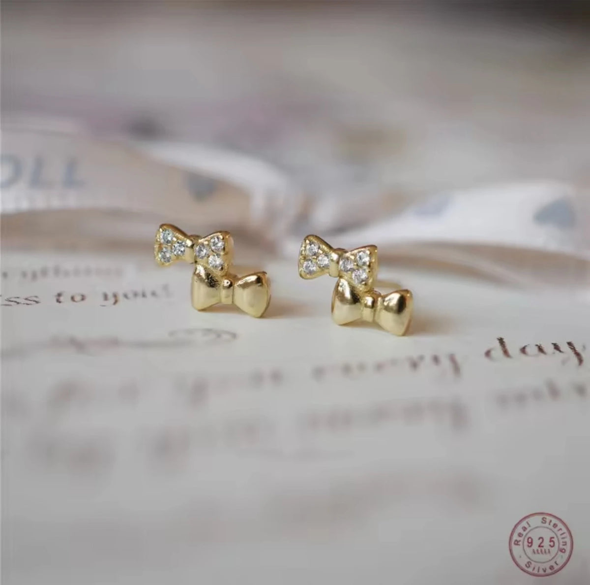A pair of gold bow stud earrings with small clear crystal accents, resting on a soft-focus beige surface with cursive text.