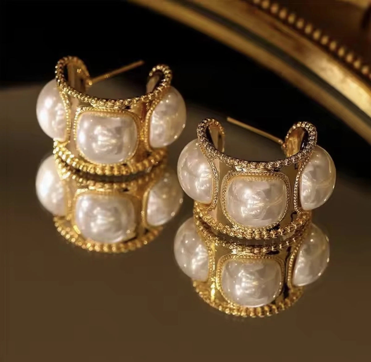 A pair of ornate gold hoop earrings with large irregular white pearl-like beads and small clear stones, displayed on a reflective dark surface with a blurred gold-toned background.